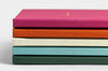 A stack of five personalized photo books in various colors, including mulberry, rust, mist, pistachio, and evergreen, all featuring a fabric texture.